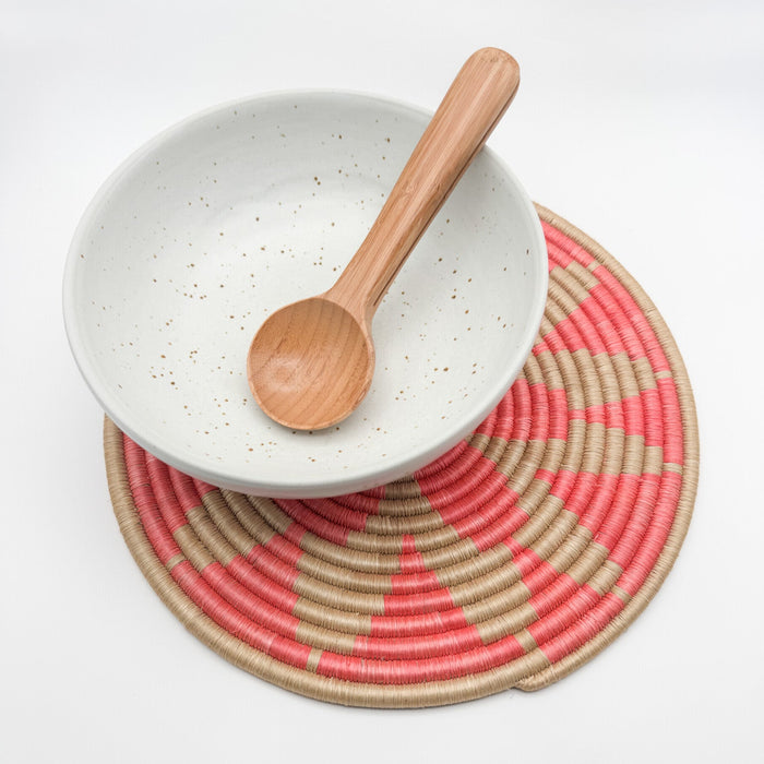 White speckled bowl with a wooden spoon on a pink and beige woven placemat.